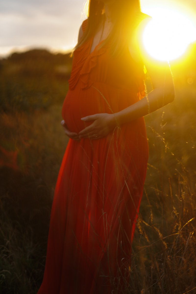 Future maman en robe rouge à contre-jour dans les herbes hautes, soleil couchant