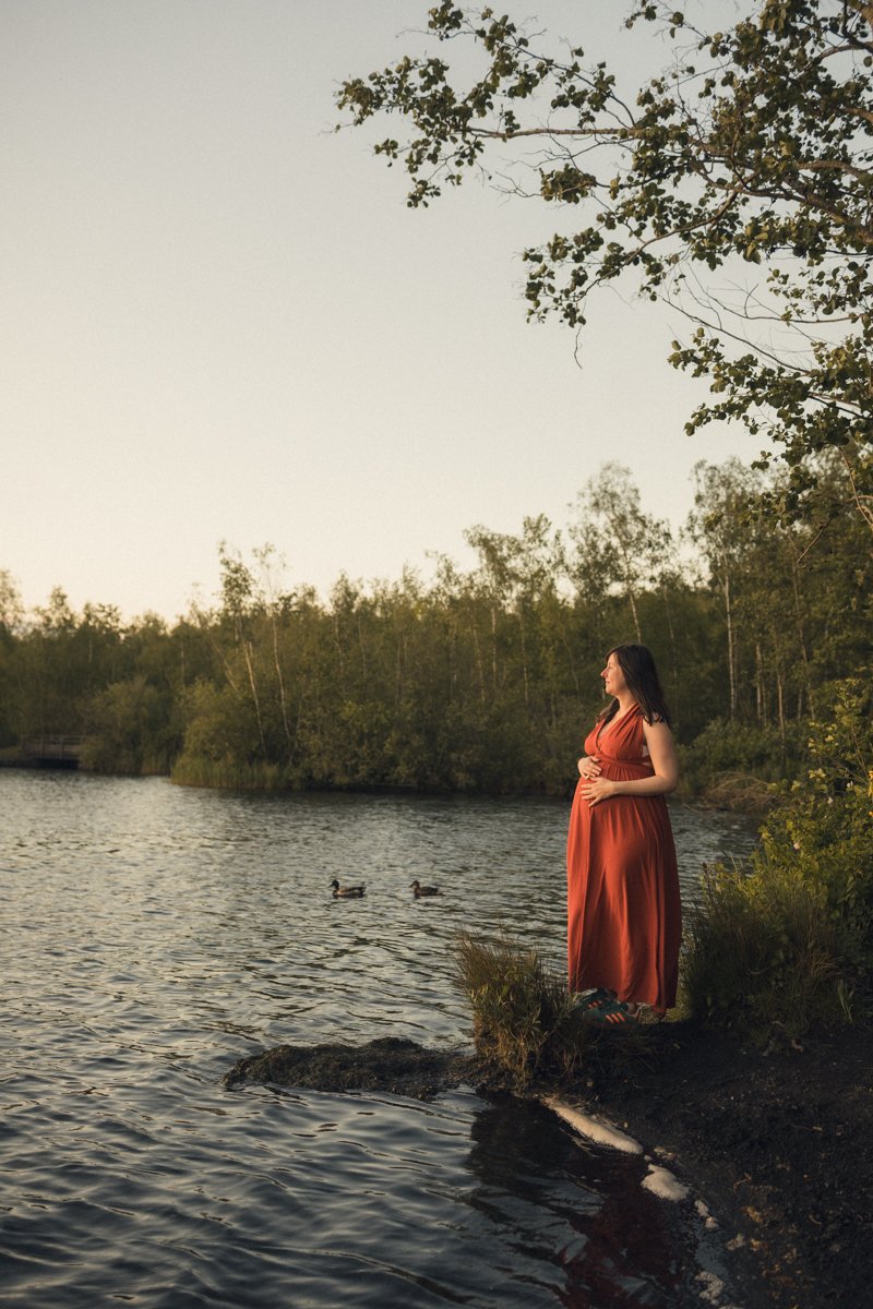 Future maman en robe longue rouge, mains sur le ventre, regardant l’étang en fin de journée
