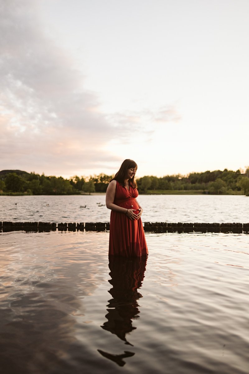 Future maman en robe rouge debout dans l’eau, son reflet sur le lac au coucher du soleil